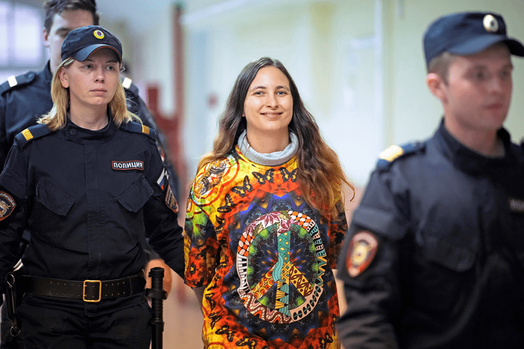 Woman with long hair wearing a bright sweatshirt with the peace sign on it, smiling, being walked by officers with hands behind her back.