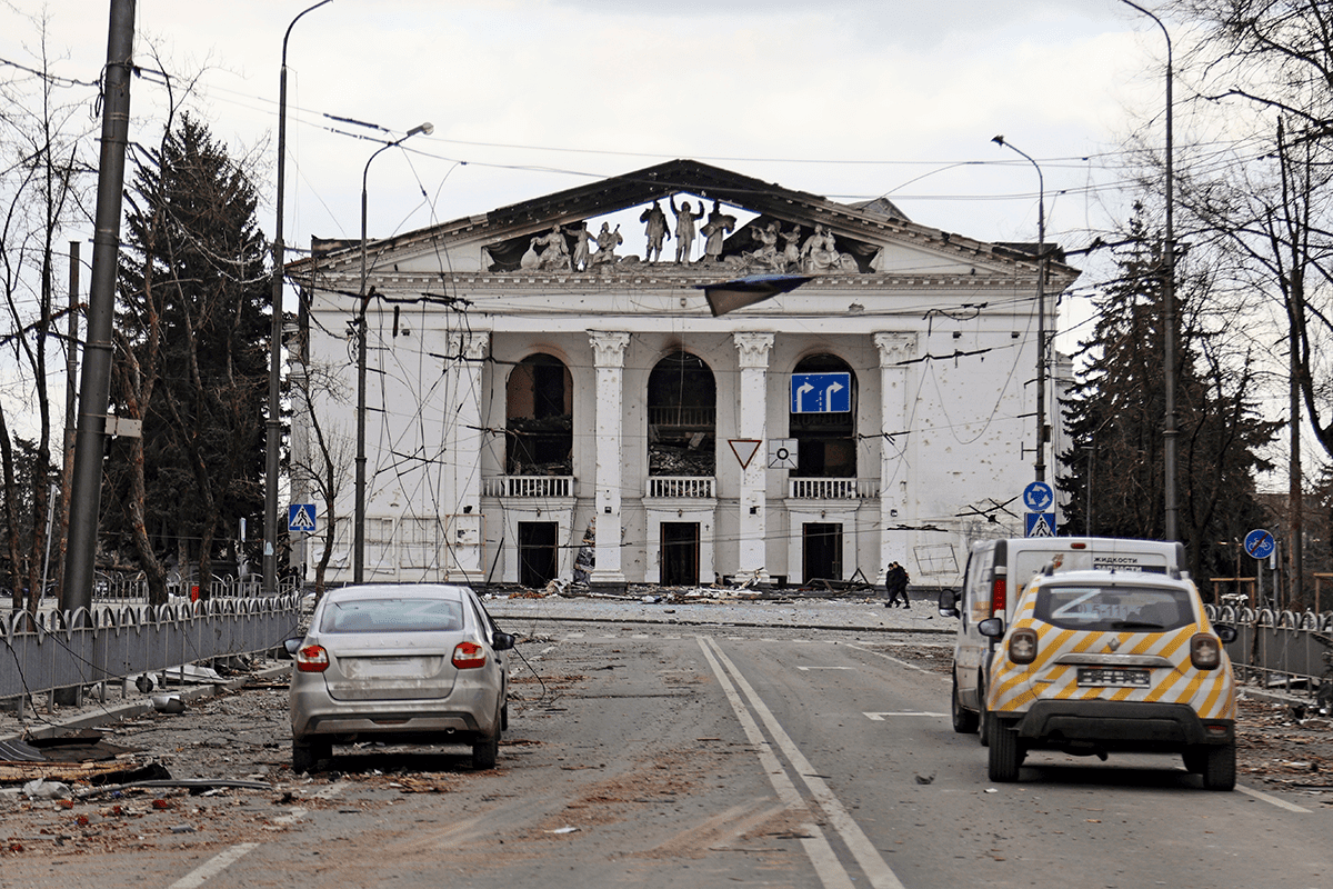 Cars on a street in front of a shattered theater building in Mariupol, Ukraine.